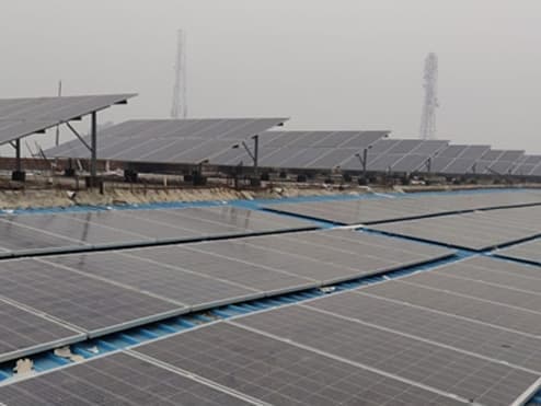 Rows of polycrystalline solar modules on a flat factory roof, with a raised row of modules behind and two lattice towers in the haze