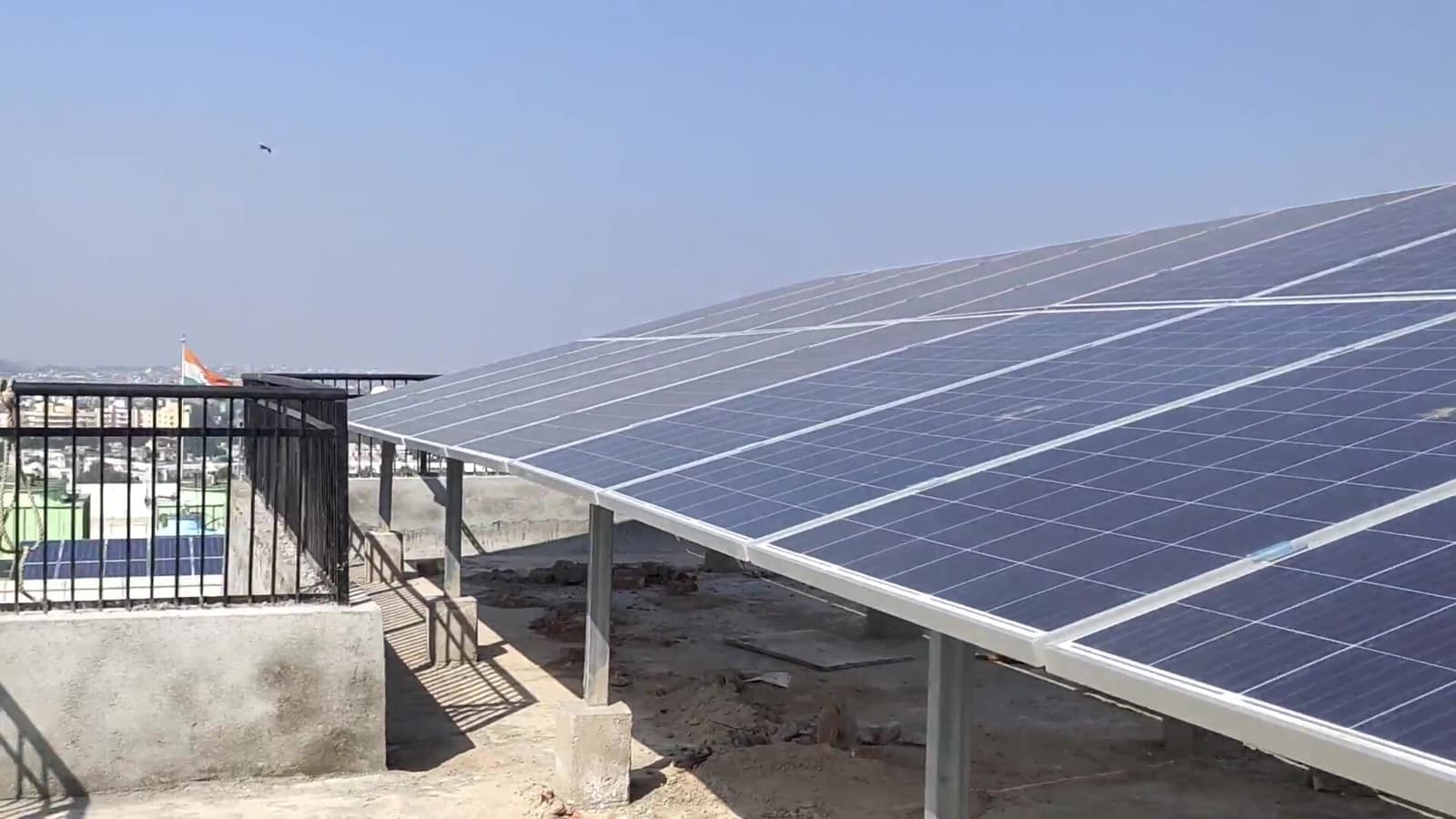 Rows of solar panels on an elevated steel structure on a rooftop, with a railing and city skyline beyond