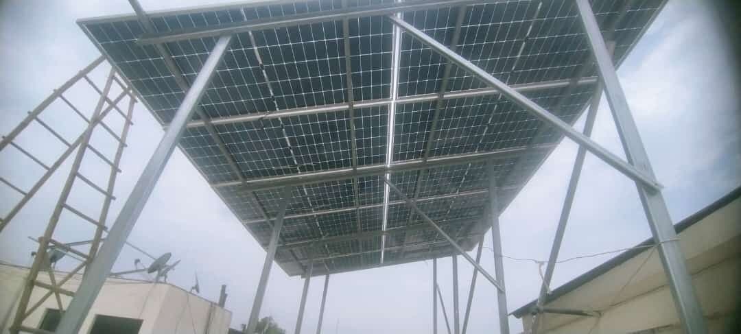 Underside of a solar panel array on a tall galvanised steel frame above a flat roof, with a ladder leaning against the structure and a satellite dish on the parapet below